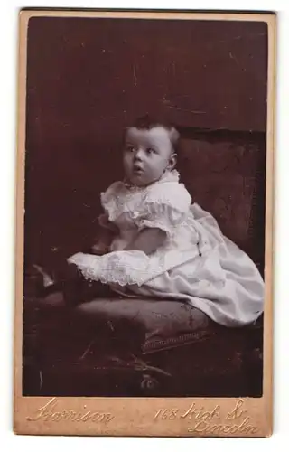 Fotografie Harrison, Lincoln, Portrait sitzendes Kleinkind im weissen Kleid mit kurzen Haaren