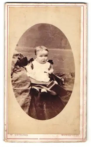 Fotografie W. Gothard, Wakefield, Portrait sitzendes Kleinkind im hübschen Kleid mit kurzen Haaren