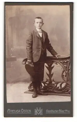 Fotografie Atelier Osten, Berlin, Junge im Anzug mit Hut stehend an einem Geländer
