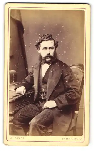 Fotografie J. Peers, Manchester, Mann mit lockiger Frisur und Vollbart im Anzug mit Fliege auf Stuhl sitzend