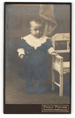 Fotografie Adolf Mewes, Rathenow, Kleiner Junge in schwarzem Mantel steht neben einem weissen Stuhl