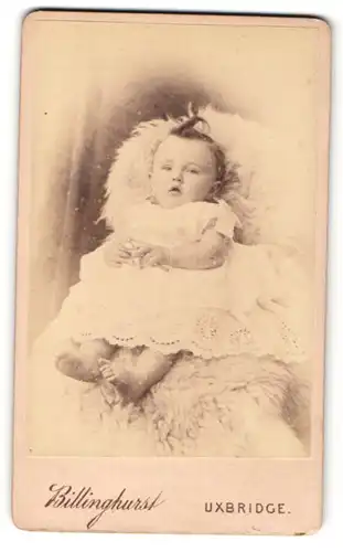 Fotografie Billinghurst, Uxbridge, Kleinkind in weissem Spitzenkleidchen sitzt auf Felldecke
