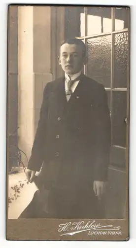 Fotografie H. Kuhlow, Lüchow i/Hann., Junger Bursche in dunklem Anzug steht neben Tisch und vor Fenster