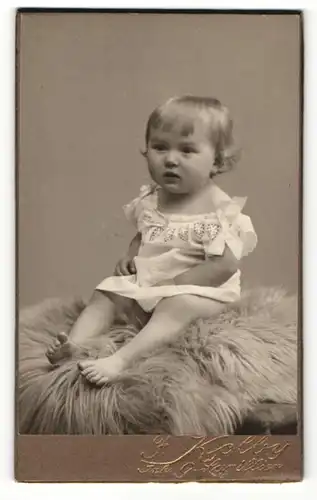 Fotografie Friedr. Kolby, Plauen, Kleines Mädchen in weissem Kleid mit Schleifen auf Lammfell sitzend