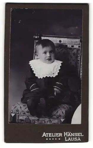 Fotografie Atelier Hänsel, Lausa, Kleinkidn in schwarzem Kleid mit weissem Zierkragen sitzt auf Stuhl