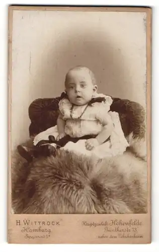 Fotografie H. Wittrock, Hamburg, Portrait niedliches Kleinkind im hübschen Kleid auf Fell sitzend