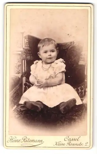Fotografie Heinr. Ritzmann, Cassel, Portrait sitzendes Kleinkind im weissen Kleid mit kurzen Haaren