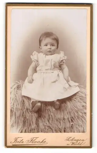 Fotografie Fritz Flaake, Solingen, Portrait niedliches Kleinkind im weissen Kleid auf Fell sitzend