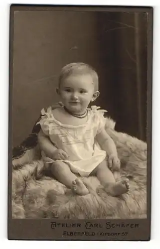 Fotografie Carl Schäfer, Elberfeld, Portrait niedliches Kleinkind im weissen Hemd mit Halskette auf Fell sitzend