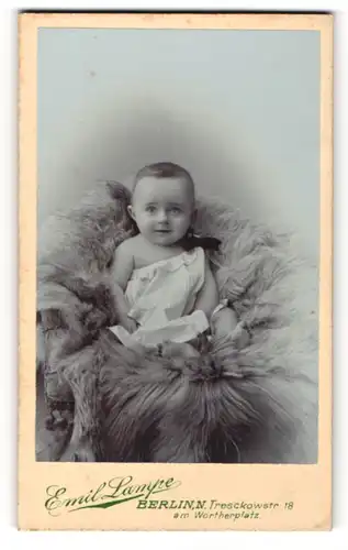 Fotografie Emil Lampe, Berlin-N, Portrait niedliches Baby im weissen Hemd auf Fell sitzend