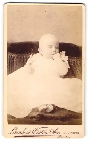 Fotografie Lambert Weston & Son, Folkestone, Portrait sitzendes Baby im weissen Kleid mit nackigen Füssen