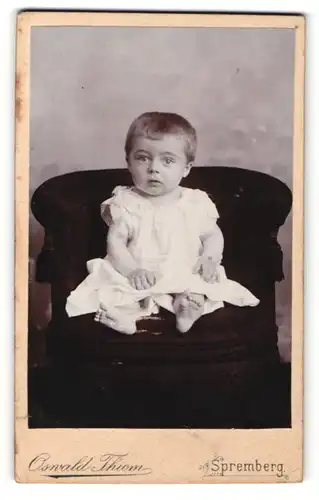 Fotografie Oswald Thiem, Spremberg, Kleinkind in weissem Kleidchen sitzt auf einem schwarzen Sessel