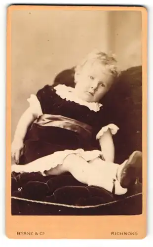 Fotografie Byrne & Co., Richmond, Kleines Kind in schwarzem Kleid mit weissen Verzierungen liegt auf Sessel