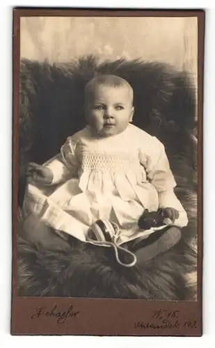 Fotografie Schaefer, unbekannter Ort, Portrait niedliches Baby im weissen Kleid mit Rassel auf Fell sitzend