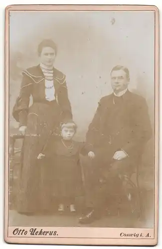 Fotografie Otto Uekers, Coswig i. A., Mann und Frau mit kleiner Tochter in schwarzem Kleid und mit Haarband