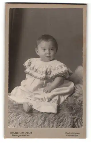 Fotografie Bruno Michaelis, Magdeburg, Portrait niedliches Kleinkind im hübschen Kleid auf Fell sitzend