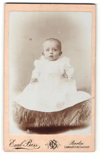 Fotografie Emil Bess, Berlin, Portrait niedliches Baby im weissen Kleid auf Fell sitzend