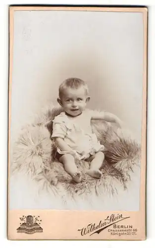 Fotografie Wilhelm Stein, Berlin, Portrait niedliches Kleinkind im weissen Hemd auf Fell sitzend
