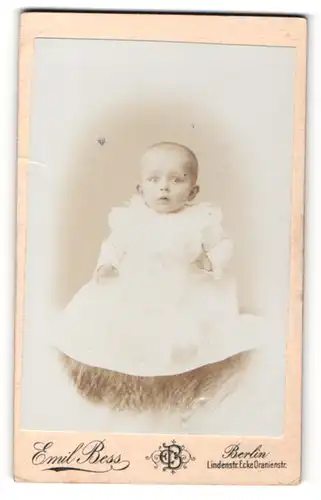 Fotografie Emil Bess, Berlin, Portrait niedliches Baby im weissen Kleid auf Fell sitzend