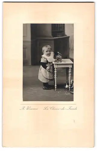 Fotografie A. Werner, La Chaux-de-Fonds, Kleines süsses Kind im Kleid greift in eine Dose auf einem Tisch