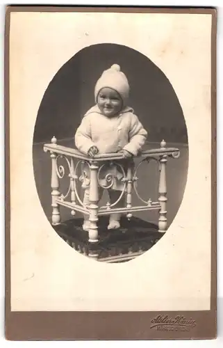 Fotografie Atelier König, Mähr. Schönberg, Kleinkind mit Mütze lehnt an einem Geländer