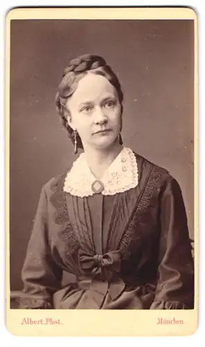 Fotografie J. Albert, München, Portrait Frau im Kleid mit weissem Spitzenkragen, elegant frisierter Zopf