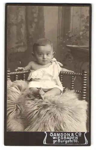Fotografie Samson & Co., Wiesbaden, Gr. Burgstrasse 10, Portrait niedliches Kleinkind im weissen Hemd auf Fell sitzend
