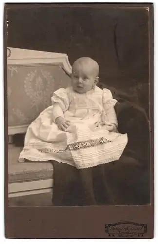 Fotografie Arthur Schmidt, Bernburg, Markt 15, Portrait niedliches Baby im weissen Kleid auf Decke sitzend
