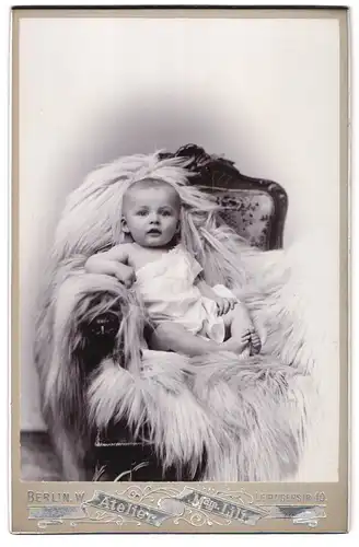 Fotografie Madame Lili, Berlin-W, Leipzigerstrasse 40, Portrait niedliches Kleinkind im weissen Hemd auf Fell sitzend