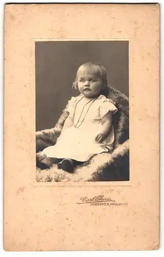 Fotografie Carl Thies, Hannover, Höltystrasse 15, Portrait niedliches Kleinkind im weissen Kleid auf Fell sitzend