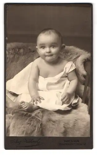 Fotografie Aug. Kühnel, Wien, Ottakringerstr. 52, Portrait süsses Baby im weissen Hemdchen auf Fell sitzend
