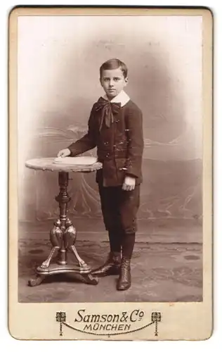 Fotografie Samson & Co., München, Neuhauserstrasse 7, hübscher Junge mit Kragenschleife stehend am Tisch