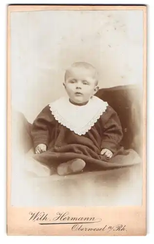 Fotografie Wilh. Hermann, Oberwesel a. Rh., Kleines süsses Kind in langem Kleid auf einem Polster sitzend