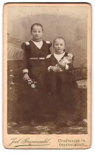 Fotografie Jul. Grunewqald, Cunewalde i. S., Portrait zwei Mädchen in Kleidern mit Puppe in der Hand
