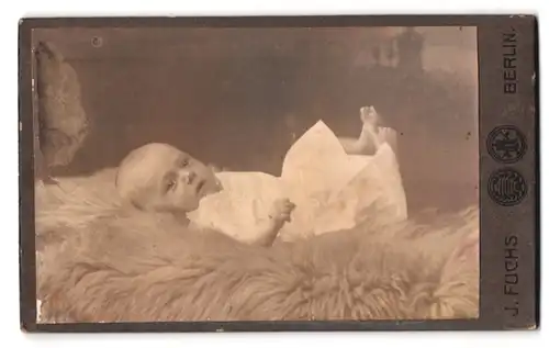 Fotografie J. Fuchs, Berlin-Neukölln, Bergstrasse 151-52, Portrait niedliches Baby im weissen Hemd auf Fell liegend