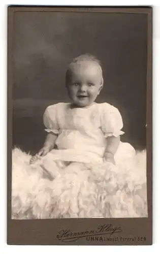 Fotografie Hermann Kluge, Unna i. Westf., Portrait niedliches Kleinkind im weissen Kleid auf Fell sitzend