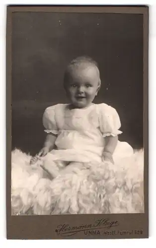 Fotografie Hermann Kluge, Unna i. Westf., Portrait niedliches Kleinkind im weissen Kleid auf Fell sitzend
