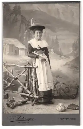 Fotografie J. Reitmayer, Tegernsee, Portrait Frau in bayrischer Tracht mit Federhut vor einer Studiokulisse