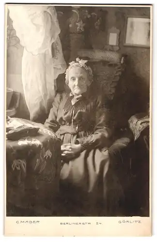 Fotografie C. Mader, Görlitz, Berlinerstr. 24, Portrait alte Frau im Kleid mit Haube sitzt in Ihrem Wohnzimmer