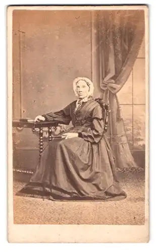 Fotografie D. Blagrove, Lewes, Portrait alte Frau im weiten Kleid mit Haube sitzend an einem Tisch