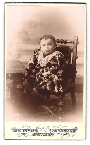Fotografie Heinr. Lutz, Kulmbach, Portrait niedliches Kleinkind im karierten Kleid am Tisch sitzend