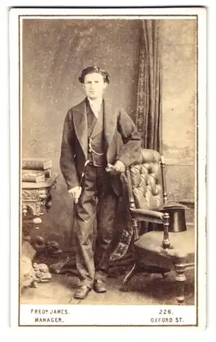 Fotografie Ferd. James, London, 226 Oxford St., Portrait Mann im Anzug mit Flanierstock und Zylinder auf dem Sessel