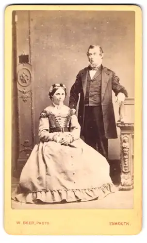Fotografie W. Beer, Exmouth, Portrait älteres Paar im Anzug mit Fliege und Sommerkleid mit Blumenhaarschmuck