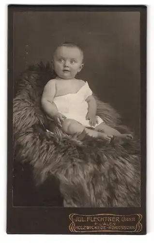 Fotografie Jul. Flechtner, Unna, Portrait niedliches Kleinkind im weissen Hemd auf Fell sitzend