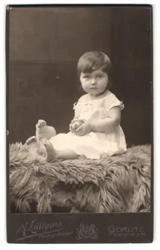 Fotografie M. Lüttgens, Görlitz, Postplatz 4, Portrait niedliches Kleinkind im weissen Kleid mit Ball auf Fell sitzend