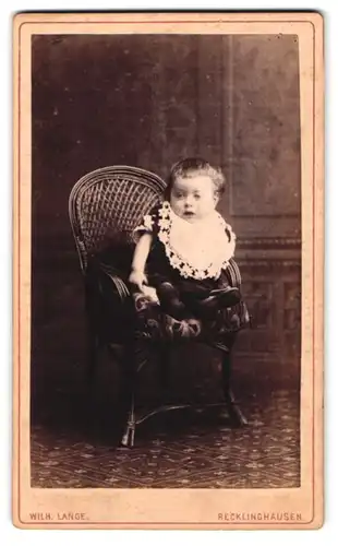 Fotografie Wilh. Lange, Recklinghausen, Kleinkind mit weissem Latz sitzt auf Stuhl mit geflochtener Rückenlehne