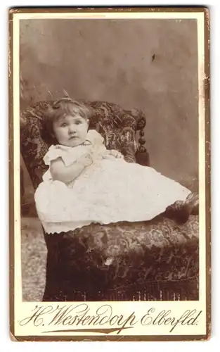 Fotografie H. Westendorf, Elberfeld, Herzog-Str. 40, Portrait kleines Mädchen im weissen kleid sitzt auf Polstersessel