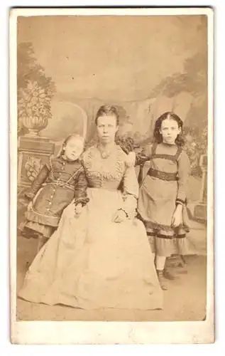 Fotografie W. New, London, 143 Brompton Road, Portrait Mutter mit zwei Kindern in Kleidern vor Studiokulisse