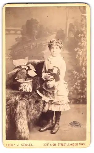 Fotografie Fred. J. Staples, Camden Town, 279 High Street, Portrait Mädchen im Rüschenkleid mit Pupp in der Hand, Locken