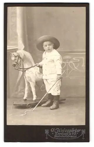 Fotografie D. Weissgärber, Zwönitz i. S., Bahnhofstr. Portrait kleiner Junge im weissen Anzug mit Schaukelpferd und Gerte